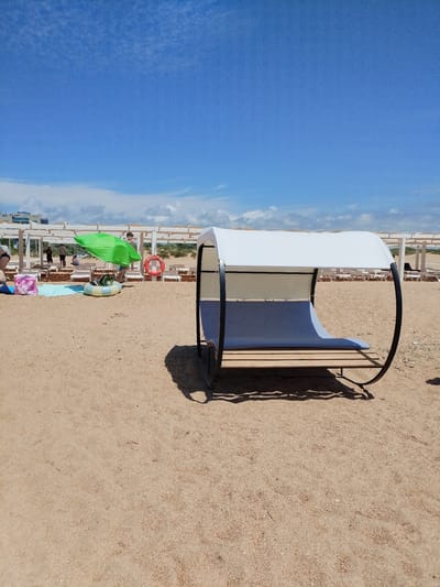The image shows a sandy beach with several rows of white lounge chairs under a pergola structure. In the foreground, there is a modern white and blue shaded lounger with a curved metal frame. To the left, a green umbrella shades some inflatable toys, beach bags, and a life preserver ring. Further back, people are visible walking and sitting. In the distance, buildings and a cloudy blue sky are visible. The location is identified as Vinogradny, Russia.