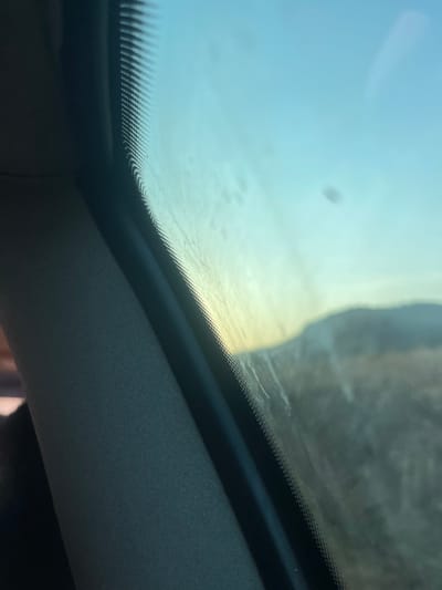 Looking upwards, the image captures the interior of a vehicle. A clear, seemingly clean sunroof is visible, with a blurry outside scene partially visible. The interior trim of the car is visible, consisting of a dark rim that goes around the sun roof and a lighter-colored upholstered roof.