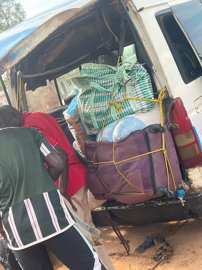 This image depicts a cargo loading scene at the rear of a white utility vehicle in Gbaza, Nigeria. The vehicle, possibly a van, exhibits significant wear and rust, particularly along its rear roofline and side panels, with exposed structural elements visible. A red and amber taillight assembly is visible on the rear passenger side, appearing partially damaged.

The rear cargo area is filled with various items, heavily secured by bright yellow ropes. Prominently visible cargo includes a large, green and white plaid patterned bag positioned at the top of the stack, a light blue bag beneath it, and a large, purple wheeled suitcase at the bottom, marked with the brand "SKYLINE." These items are tightly packed and crisscrossed with multiple strands of yellow rope, indicating a method of securing the load.

Two adult males with dark complexions are present. In the foreground on the left, a man is seen from the back, wearing a dark green athletic top featuring a distinctive lower panel with vertical white stripes interspersed with thin red lines, on a dark background. He is also wearing dark pants with a light vertical stripe. He appears to be leaning towards the vehicle. Partially obscured by the first man and the cargo, a second man in a red collared shirt and light-colored trousers is leaning into the vehicle's cargo space. Both individuals appear engaged in the process of loading or securing the items.

The immediate ground surface is unpaved, consisting of light brown or tan dirt. A tangled bundle of various ropes or straps lies on the ground directly below the vehicle's rear bumper. The background is blurred, showing distant foliage and light-colored building structures under a bright, overcast sky.