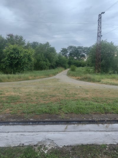 The image depicts a rural landscape with a dirt road that forks into two paths, surrounded by lush green trees and grass. In the background, a rusty metal pole with electrical wires stretches towards the cloudy sky, and a faint structure is visible in the distance. The foreground shows a concrete structure with metal rails, possibly part of a railway line, bordering the grassy area. The scene appears to be an undeveloped area, likely on the outskirts of a town or city. The weather is overcast, suggesting a cloudy or possibly rainy day. No people or animals are visible in the frame. The overall impression is one of natural simplicity and quietude.