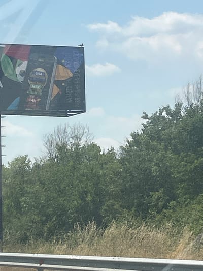 The image captures a billboard against a sky with scattered clouds. The billboard features graphics that appear to represent various national flags or country names like "USA", "MEX", and "CAN". To the right of these country-related graphics, there is text in an alphabet that is not Latin. The billboard is positioned above a dense line of green trees and dry grass, with a metal guardrail visible in the foreground. A small, dark object, possibly a bird, is perched on the top edge of the billboard. The scene suggests an outdoor setting, likely along a road, given the presence of the guardrail.