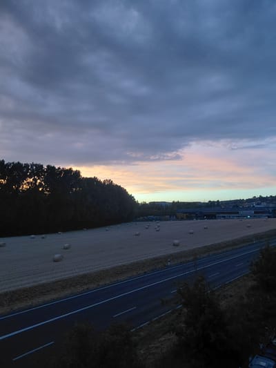 The image depicts a rural landscape near Ecublens, Switzerland, at dusk. In the foreground, a two-lane road with white markings runs diagonally from the bottom left to the bottom right. Adjacent to the road, on the left, is a field with numerous round bales of hay scattered across it. A dark line of trees runs along the far edge of the field, separating it from the horizon.
In the mid-ground, a low-rise industrial or commercial building complex is visible. It features a light-colored facade with dark windows and signage that appears to read "boissec". Beyond the buildings, a slightly undulating hillside is covered with scattered houses and more trees, forming a distant treeline against the sky.
The sky is predominantly filled with dramatic, dark gray clouds, especially in the upper portion of the image. Towards the horizon, there is a soft gradient of light, with hues of pale orange, pink, and a pale greenish-blue suggesting the last light of sunset or the first light of dawn. The overall impression is of a twilight scene over a cultivated agricultural area bordering a developed commercial zone. There are no people visible in the image.