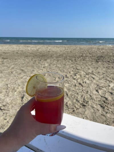 The image shows a close-up of a hand holding a clear glass filled with a red beverage and garnished with a slice of lemon. The hand has light-colored fingernails. The glass is partially obscured by the hand and is positioned over a sandy beach. In the background, the ocean and the sky are visible, suggesting a daytime setting at a beach location. The sand appears to be fine-grained and light brown. To the right of the glass, a white surface, possibly part of a lounge chair, is visible. The lighting suggests a bright, sunny day. The overall mood conveyed is one of relaxation and leisure, typical of a beach vacation. The location context provided is Skala Avdiron, Greece. No text is visible in the image.