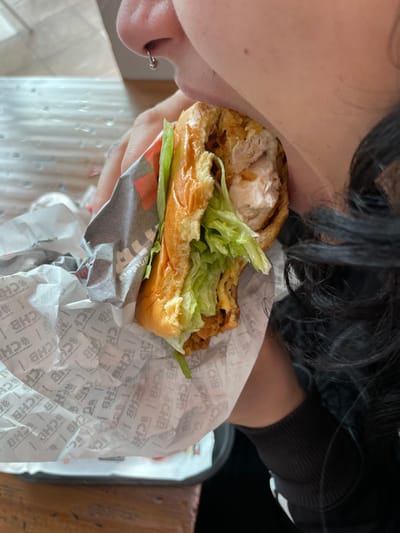 A close-up image depicts an individual in Plaisir, France, in the process of consuming a chicken burger. The person's lower face, including the nose with a small silver hoop piercing in the right nostril, and part of their mouth are visible on the upper right. Dark, curly hair frames the right side of the image, and a sleeve of a dark, ribbed garment is present. The individual's left hand holds a partially eaten chicken burger wrapped in white paper branded with repeating "#CHB" text. The burger consists of a golden-brown bun, a substantial piece of fried chicken with exposed white meat, and green lettuce. A black plastic tray rests on a wooden table surface in the lower left, indicating an indoor dining environment.