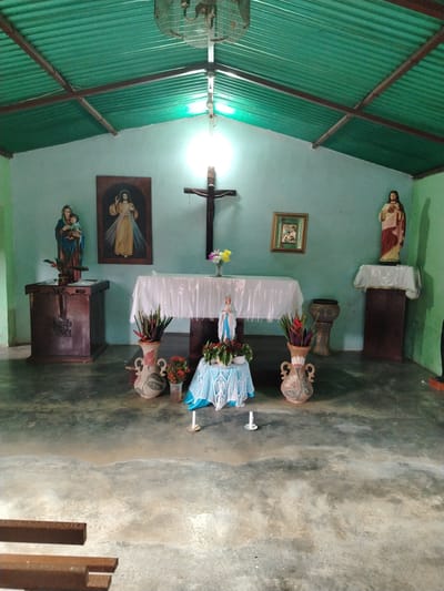 The image depicts a small, simple chapel in what appears to be Los Leoncitos, Venezuela. The building has a corrugated metal roof supported by exposed wooden beams. The walls are painted a light blue-green color.
Inside, the chapel contains several religious objects. On the left is a statue of Mary holding the infant Jesus atop a wooden pedestal. Next to it hangs a framed picture of Jesus. In the center, a table covered with a white cloth holds a crucifix and a vase of flowers. Below the table is a smaller table draped with a light blue crocheted cloth, topped with a statue of Mary surrounded by flowers. Two candles stand on the floor in front. Vases containing flowers flank this setup. On the right side of the chapel, there's a statue of Jesus atop a wooden pedestal. A framed picture also hangs on the wall. A drum sits between the alter table and the statue.
The floor is concrete and appears to be well-worn and possibly damp. A few wooden bench slats are visible in the foreground. The lighting is provided by an overhead light fixture, resulting in a bright spot on the back wall behind the crucifix. The overall impression is of a modest, well-used, and cared-for religious space.