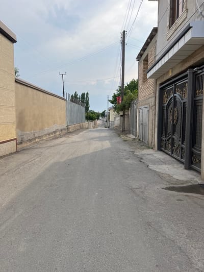 The image shows a narrow, asphalt road in a residential area, likely in the city of Guliston, Tajikistan. The road is lined with walls and buildings on both sides. On the left, there is a tan-colored wall with a textured finish, followed by a corrugated metal fence. On the right, a building with a light-colored facade is visible, featuring a large, ornate black gate with gold detailing. Further down the street, more buildings and fences can be seen, along with trees and utility poles with power lines. The sky is partly cloudy. The overall impression is of a quiet, somewhat worn urban streetscape.