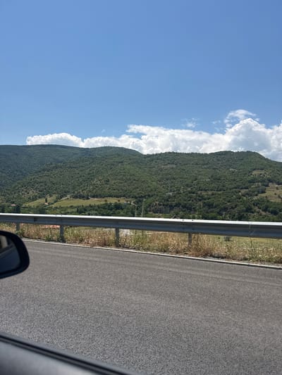 The image presents a panoramic view of a mountainous landscape from the perspective of a vehicle, likely taken in Jeleznitsa, Bulgaria. The scene is dominated by a clear blue sky and lush green mountains.

In the foreground, a dark asphalt road occupies the right side of the frame, indicating the direction of travel. A silver metal guardrail runs parallel to the road, separating it from a strip of dry, light-colored grass and weeds. A portion of a car's side mirror is visible in the bottom right corner, reinforcing the viewpoint from a moving vehicle.

The midground and background feature a majestic mountain range, heavily covered with dense green trees and foliage. Patches of terraced land or clearings are discernible on the mountain slopes, along with a few scattered small buildings, possibly houses or farm structures. Faint lines, possibly power lines, can be seen traversing parts of the mountainside.

The sky above the mountains is a vibrant blue, filled with large, fluffy white cumulus clouds that appear to rest atop the mountain peaks. The bright natural light suggests it is daytime, likely a sunny morning or afternoon. There are no people visible in the image, nor any discernible text. The overall atmosphere is one of serene natural beauty and travel through a picturesque landscape.