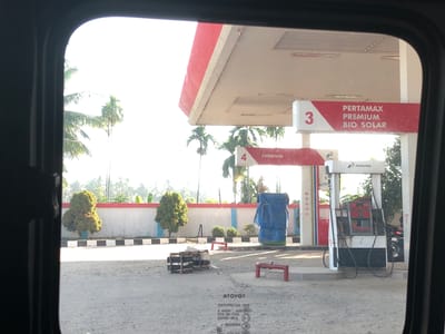 The image captures a bright, sunny view of a Pertamina gas station (SPBU) in Kutalawah, Indonesia, seen from inside a vehicle through its window. The dark frame of the window occupies the left and right edges, with a rounded top edge.
The gas station features a red and white canopy structure, with two distinct pump islands visible. On the right, a sign under the canopy reads "PERTAMAX PREMIUM BIO SOLAR" next to the number "3". Further left, another sign indicates "PREMIUM" next to the number "4". A gas pump with "PERTAMINA" branding is prominently displayed on the right side of the frame, showing its hoses and nozzles. A motorcycle is partially visible behind this pump.
The ground around the station is a light-colored, unpaved surface, possibly gravel or packed dirt. In the background, numerous palm trees and other lush green foliage are visible under a bright, almost white sky, suggesting strong daylight, likely morning or late afternoon given the clear shadows. A low wall with a black and white checkered curb runs across the mid-ground, separating the station area from the greener landscape. Behind this wall, some small, manicured bushes and larger trees grow.
Several objects are scattered on the ground: two red, low-lying benches, and a stack of what appears to be wooden crates or barrels, and a large object covered with a blue tarp.
Reflected on the car window glass in the foreground, faint, reversed text can be seen, including "ATOYOT" (likely "TOYOTA" backwards) and other alphanumeric sequences, suggesting reflections from the vehicle's interior. No people are explicitly visible in the scene.