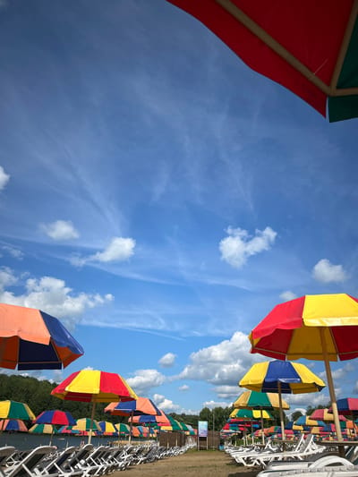 The image depicts an outdoor scene at a beach or recreational area, likely in the city of Yubileiny, Russia, given the prompt's reference. The foreground is dominated by rows of white lounge chairs arranged on sandy ground. Behind the chairs, a multitude of colorful beach umbrellas are set up. These umbrellas are multi-colored, featuring combinations of red, yellow, blue, and green panels.

In the background, a body of water, possibly a lake or river, is visible, bordered by a line of trees. The sky above is a clear blue, punctuated by scattered white clouds, some of which are wispy and others more substantial. The overall impression is one of a bright, sunny day at a leisure facility, prepared for visitors. No people are clearly visible in the image.