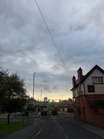 The image captures a street scene in Oswestry, United Kingdom. The sky is overcast with varying shades of gray clouds. On the right side of the frame, a red brick building with a sign reading "The Plough" is visible. A road curves into the distance, marked with double yellow lines on the edge. A petrol station stands further down the road, with green and white signage. A tree with sparse foliage is situated in the foreground. A street light pole is visible between the tree and the petrol station. A street sign with a red circle and white arrow indicating "no right turn" is also present. A thin wire stretches across the sky from the building on the right towards the upper left corner of the image.