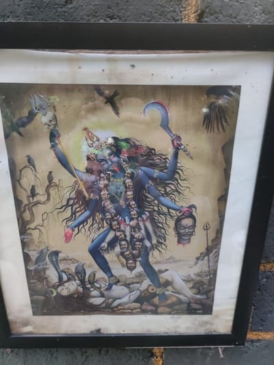 The image is a framed print depicting the Hindu goddess Kali standing on a prone figure of Shiva. Kali has multiple arms, holding a severed head, a sword, and a trident with a skull on top. She wears a garland of severed heads. The print features crows and other birds in the background. The frame is black. No information is available to verify this image's location in the city of Bedulu, Indonesia.