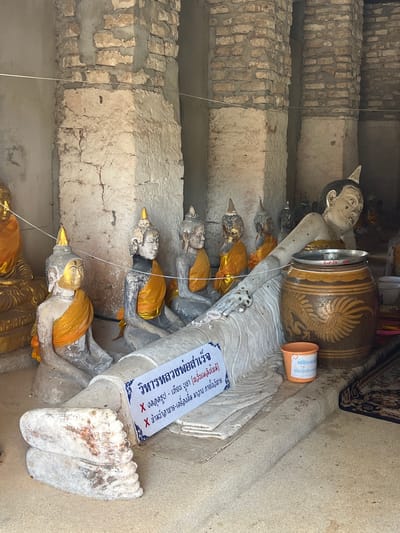 The image shows the interior of a building at Baan Tha Po, Thailand, featuring several statues of the Buddha. In the foreground, a large reclining Buddha statue is prominently displayed, lying on its side with its head resting on its arm. To the left of the reclining Buddha, a seated Buddha statue is visible, adorned with orange cloth. Several other seated Buddha statues, also draped in orange cloth, are positioned behind the reclining figure. The statues are made of stone or plaster and appear to be weathered. A large ceramic pot with decorative markings is placed next to the reclining Buddha. A sign with Thai text is positioned in front of the reclining Buddha. The environment is rustic, with exposed brick pillars and a concrete floor. The lighting suggests an indoor setting, possibly a temple or historical site.