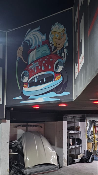 This media file displays a mural on a dark blue wall depicting a cartoon car being washed. A hand holding a yellow sponge is seen washing the front of a red and blue car. Water splashes are depicted around the car, along with a hose being used to spray water. The car has large, round headlights and a stylized grille. Red LED lights are visible along the edge of the wall, suggesting an indoor setting, possibly a garage or workshop. In the background, to the right, shelves filled with car parts are visible, indicating an automotive-related location. The overall style of the mural is playful and cartoonish. Text is partially visible on the top wall, appearing to be "FT GARAGE" in white letters with a red stripe. The image is rotated 90 degrees counter-clockwise.