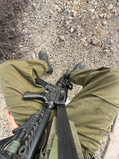 A person is sitting on a rocky surface with a black rifle lying across their lap. They are wearing green military-style pants and black boots. The rifle is held in place with a black strap around the person's waist. The location is a dry, rocky area near the city of Mazkeret Batya, Israel. The image is taken from a low angle, looking up at the person and the rifle. It is difficult to determine the person's gender or age based on the image. The person is not interacting with anyone or anything in the image.