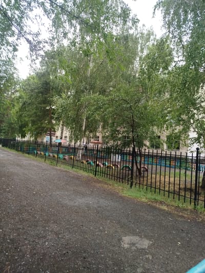 The image depicts an outdoor scene, likely a schoolyard or park in Kabanbay, Kazakhstan. The foreground is dominated by a gravel path that curves to the left, suggesting a pedestrian walkway. To the right of the path, a black metal fence encloses a grassy area with a playground. The playground features colorful elements, possibly made from repurposed tires, and some scattered pieces of playground equipment. Beyond the fence and playground, a building with multiple windows and a light-colored facade is visible, partially obscured by lush green trees. The trees are a mix of deciduous and coniferous species, with dense foliage suggesting it's summertime or late spring. The sky appears overcast, indicating a cloudy day. No people are clearly visible, but the presence of the playground suggests it is a place for children. The overall mood of the image is calm and perhaps a little subdued due to the weather. There is no visible text in the image.