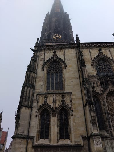 The image displays the St. Lamberti Church (St. Lamberti-Kirche) in Münster, Germany, from a low-angle perspective. The gothic stone facade and its towering, dark spire dominate the frame, extending upwards into an overcast or foggy sky.
The church's architecture is characterized by light-colored weathered stone, dark-paned pointed arched windows with intricate tracery, and decorative sculptural elements including gargoyles. A prominent golden-faced clock is visible on the spire, indicating a time of approximately 11:10. Above the clock, the dark, tapering spire features multiple narrow arches and culminates in a sharp point. The uppermost section of the spire, partially obscured by the atmospheric haze, clearly exhibits three distinct, dark, horizontal cage-like structures.
In the lower-left corner, a small section of an adjacent building with a red-tiled roof is partially visible. No individuals or discernible activities are present within the scene. The overall lighting suggests a uniformly diffused, non-sunny environment.