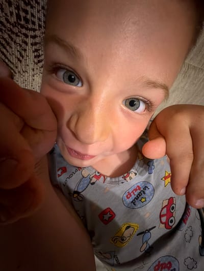 This is a close-up, low-angle indoor photograph of a young child lying down. The child has light skin, light eyebrows, and gray-green eyes. Their mouth is slightly open, and their tongue is visible. The child is wearing a gray t-shirt with various cartoonish prints, including a red car with big eyes, a cartoon airplane, clouds, and text that says "island trip". There is also a character that resembles Snoopy from the Peanuts comic strip. The child's hands are visible in the foreground and near their face. The background appears to be a textured surface, possibly a rug or carpet, with a dark pattern. The lighting suggests an indoor setting, potentially in the evening or on an overcast day, with a soft, diffused light source. The overall mood of the image is intimate and candid, capturing a moment of quiet play or rest. The location context provided is Zmeitsa, Bulgaria, but there are no specific visual cues in the image to confirm this location.