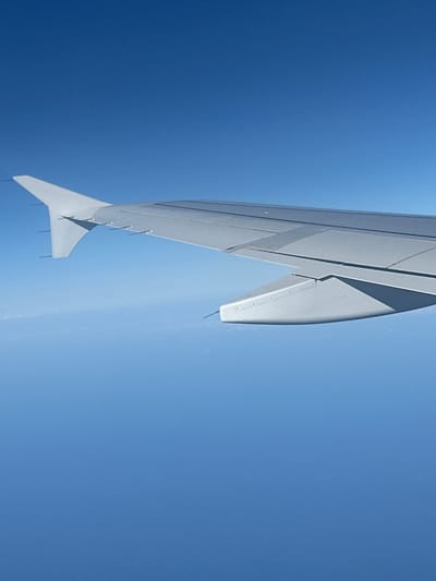 The image shows the underside of an airplane wing against a clear blue sky. The wing is a light gray color and has a sleek, aerodynamic design with visible panel lines and a winglet at the tip. The wing occupies the left and central portion of the frame, angled downwards from the top left corner. The sky is a uniform, bright blue, suggesting it is daytime. There are no visible clouds, indicating fair weather. The perspective suggests the viewer is inside another aircraft, looking out from a window, or is positioned below and to the side of the wing. The scene conveys a sense of travel and being airborne. No people, text, or specific location cues are visible, though the provided location context is Yehud-Monosson, Israel.