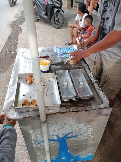 A street vendor prepares food on a metal cart featuring a hand-painted blue tree. The cart holds trays of filled buns, containers of toppings, and bags of ingredients. A vendor in a t-shirt and cargo shorts is cutting buns. In the background, a woman and child sit near a parked motorcycle on a sidewalk. A person wearing a gray sweatshirt is also present.