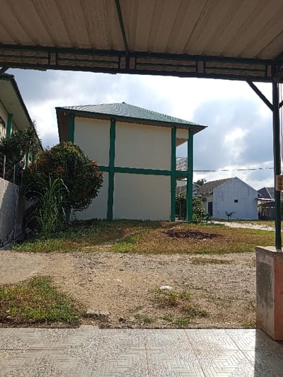 The image depicts an outdoor scene in Keudematangglumpangdua, Indonesia, viewed from beneath a light-colored corrugated metal canopy supported by dark metal framework. In the immediate foreground, a patterned tiled surface is visible, extending to the bottom of the frame. A concrete pillar is partially visible on the right. The midground consists of an unpaved area of earth, sparsely covered with green grass and patches of dry, reddish-brown vegetation.
In the background, a two-story building with cream-colored walls and prominent green structural elements stands on the left. It features a dark green corrugated hip roof. A large, rounded green shrub with some reddish foliage is situated in front of this building, accompanied by taller green plants. Further to the right, a smaller, single-story white building with a dark roof is visible. The sky is overcast with a mix of darker clouds and brighter areas. No individuals are present in the scene.