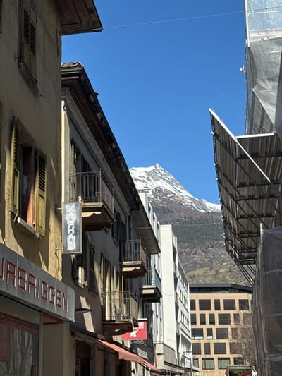 The image depicts a street view in Visp, Switzerland, under a clear blue sky. On the left, an older, multi-story building with a light beige facade features balconies with dark metal railings and numerous closed louvered shutters, some appearing worn. A vertical, dark-colored sign with light, stylized characters hangs from this building. Below it, the red signage "URBRIGGEN" is partially visible. Further down the street, a red sign with a white elephant and the word "THAI" is affixed to another building. The right side of the scene is framed by extensive scaffolding covered in protective white sheeting, indicating ongoing construction or renovation. In the midground, modern, light-colored buildings are visible. In the background, a prominent snow-capped mountain peak rises under the bright blue sky, with its lower slopes showing sparse vegetation. A thin wire runs horizontally across the upper portion of the frame. No individuals are clearly discernible.