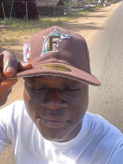 A close-up selfie depicts a man wearing a brown baseball cap with an embroidered logo of what appears to be a marlin and an "L," alongside a white t-shirt. The man is holding the brim of the cap. The background suggests an outdoor location, possibly a street, with vegetation and structures visible in the distance. Based on the prompt, the location is Ikoga, Nigeria. The environment includes a road and what appears to be some residential or commercial buildings. The light suggests a sunny day.