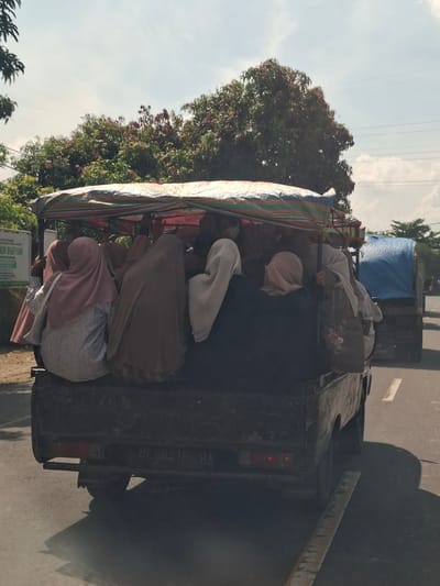 In Gampongrayadagang, Indonesia, a group of people are being transported in the back of a pickup truck modified with a covered canopy. The truck bed is filled with individuals, primarily women wearing hijabs, seated closely together. The canopy appears to be made of striped fabric and is supported by poles. Some people are looking forward, while others are looking sideways or down. In the background, lush green trees line the side of the road under a partly cloudy sky. Power lines are visible overhead. The truck is driving on a paved road, with a white line marking the lane. Further down the road, another truck covered with a blue tarp is also visible. The license plate of the pickup truck reads "BL 8416 BA".