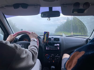 The image shows the interior of a car from the perspective of the driver. The driver's hands are visible on a pink-covered steering wheel. A smartphone mounted on the dashboard displays a navigation app. The dashboard also shows a car radio, air conditioning controls, and a gear shift. Outside the car, a tree-lined road can be seen under a cloudy sky. The scene appears to be a road trip in or near Khomutovka, Russia, as indicated by the location context. The weather appears overcast, possibly raining, with water droplets visible on the windshield. The interior of the car is functional and appears to be in moderate condition. No other people are clearly visible in the car, although a person's legs and arm are visible on the passenger side, suggesting at least one other occupant.
