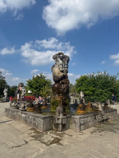 In Cambron-Casteau, Belgium, a large stone fountain stands as the central feature of an outdoor plaza. The fountain itself is a multi-tiered sculpture of stacked, anthropomorphic figures, with water cascading down its sides. Around the base of the fountain, a square, stone-lined pool is adorned with smaller sculptures, including several elephant figures, and ornate, conical structures that appear to be spouting water. The plaza is paved with large, irregular stone slabs, some of which are wet, suggesting recent water activity.

To the left of the fountain, a red bridge can be seen, with several people seated and standing on or near it. Further left, a lighthouse-like structure and a statue of a standing figure are visible. Behind the fountain and to the right, lush green trees and foliage provide a natural backdrop. In the distance, a tall industrial chimney with red and white bands is present.

On the right side of the frame, a person in a dark jacket and light-colored pants stands, appearing to be looking at their phone. The sky above is a bright blue, dotted with fluffy white clouds. The overall impression is of an outdoor decorative installation, likely a park or garden attraction, with a mix of artistic sculptures and natural elements.