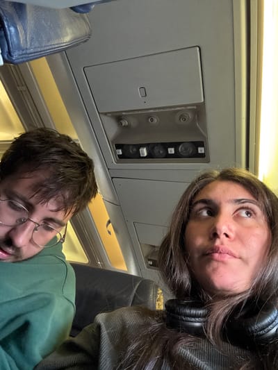 The scene is an interior view of an aircraft cabin, located in the city of Tzafria, Israel. Two individuals occupy seats in the cabin. On the left, a male with dark hair and gold-rimmed glasses is depicted with his head tilted to his right, wearing a green hooded garment, and appearing to be asleep or resting. On the right, a female with long dark hair and brown eyes is looking upwards and to her right. Black over-ear headphones are resting around her neck over a grey, ribbed top. The overhead cabin panel is visible, featuring air vents, reading lights, and call buttons. A partially visible blue-grey overhead bin is in the upper left. Yellowish light from a window or cabin lighting illuminates the right side of the frame and the background. The seating appears to be dark-colored.