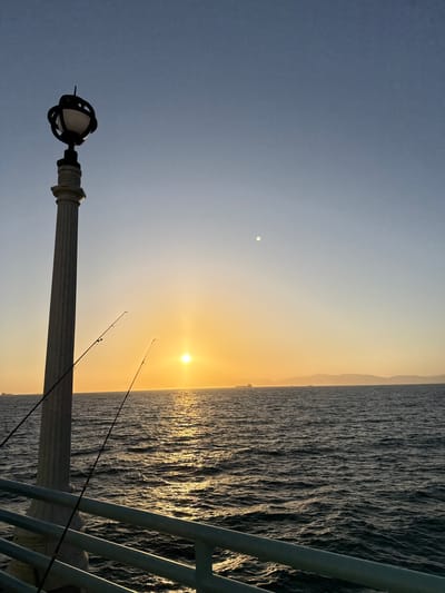 This is a vertical, eye-level, full-shot photograph of a sunset over the ocean from a pier in Manhattan Beach, California. In the foreground, a decorative lamppost with a white column and a black, ornate lamp fixture is positioned on the left side of the frame. Two fishing rods are leaning against the lamppost, with their lines extending towards the water. A pale green railing runs along the bottom of the frame. The ocean fills the middle ground, with moderate waves reflecting the warm hues of the sunset. In the background, a hazy shoreline with hills and mountains is visible, with a few ships or boats dotting the horizon. The sun, a bright orange orb, is near the center of the frame, casting a shimmering golden path across the water. The sky transitions from a pale yellow near the horizon to a clear, soft blue at the top of the frame, with a faint white circle, possibly the moon, visible in the upper portion of the sky. The overall mood is serene and tranquil.