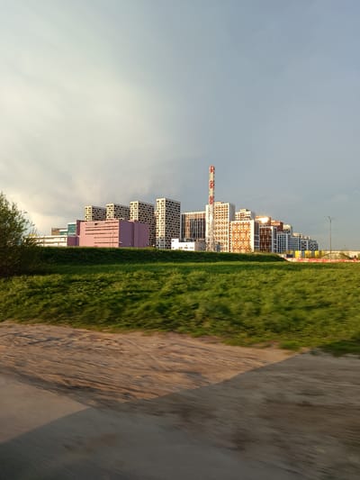 The image depicts a cityscape in Novoivanovskoe, Russia, viewed from a slightly elevated position across a grassy expanse. A cluster of modern, multi-story buildings dominates the horizon. The buildings are varied in architectural style and color, including structures in white, beige, and a distinctive pink hue. A tall chimney, marked with red and white stripes, stands prominently among the buildings. The foreground is dominated by green grass and an area of sandy or unpaved ground. The sky is overcast with a mix of light and dark clouds. The image exhibits motion blur, suggesting it was taken from a moving vehicle. There are no people visible in the scene.
