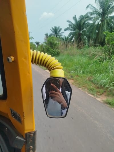 The image depicts a view from inside a yellow vehicle, possibly an auto-rickshaw, moving on a paved road. The road is flanked by green vegetation, including tall grasses and palm trees, characteristic of Ukwulu, Nigeria. The vehicle's side mirror reflects a person holding a smartphone, presumably taking the picture. The sky appears overcast.