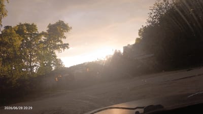 The image depicts a view of the sky and a rural landscape at dusk, possibly during sunset, with visible sun rays. In the foreground, a dirt road or path is visible, with the hood of a vehicle partially obstructing the lower portion of the frame. To the left, lush green trees with sunlight filtering through them are prominent. Further into the scene, several low-rise buildings, likely houses or farm structures, are scattered amongst the trees. A line of coniferous trees forms a backdrop on a distant hill. The sky is a pale orange and yellow, with clouds diffused by the bright sunlight. The timestamp at the bottom left corner indicates the date as June 28, 2026, at 20:29. The scene suggests a serene, possibly remote, countryside setting in or near the city of Luza, Russia.