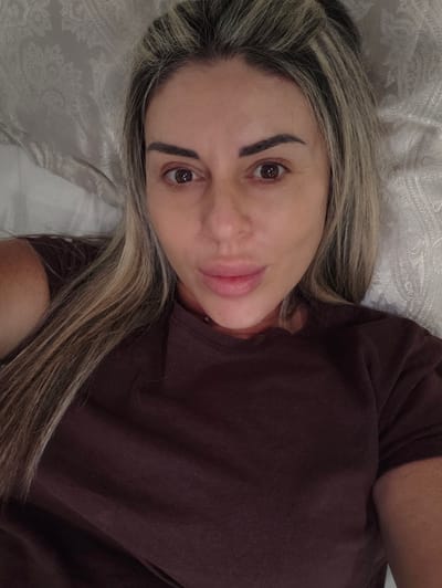 A medium shot portrait of a woman lying down, looking directly at the camera. She has long, blonde-streaked brown hair that is partially pulled back. Her skin is fair, and she is wearing a dark brown t-shirt. The background appears to be a bed with textured white sheets. The image does not contain any information that would allow for the determination of its location in Carpi, Italy.