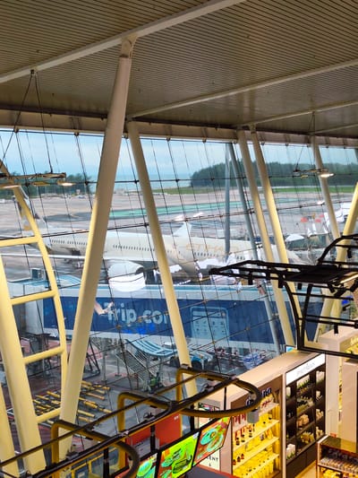 The image depicts the interior of an airport terminal in Tambon Mai Khao, Thailand, looking out onto the tarmac. The terminal features a modern architectural design with large glass windows supported by a network of white and yellow structural beams. Through the glass, several aircraft are visible parked on the runway. One prominent aircraft, identified by the "ETIHAD" logo, is partially obscured by the terminal's structure. The tarmac area shows various airport operational elements, including jet bridges, service vehicles, and what appear to be luggage carts. In the foreground, within the terminal, a retail store selling skincare and cosmetic products is visible, featuring well-lit shelves displaying numerous bottles and containers. Digital screens displaying food items and promotions are also present in the retail area. The environment outside the terminal suggests a cloudy or overcast sky over a body of water and a treeline.