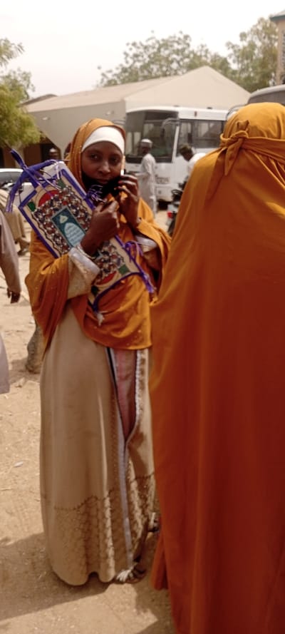 This image depicts an outdoor scene in Sani Mai Nagge, Nigeria, characterized by a dusty, unpaved ground surface. The primary subject is a woman in the mid-ground, facing slightly towards the viewer. She wears a mustard yellow headscarf over a white undercap, a matching long-sleeved garment with subtle reflective elements, and a light beige long skirt featuring patterned vertical and horizontal strips. Her right hand is raised, holding a dark object, possibly a phone, near her ear/mouth. She carries a rectangular, decorative object with blue trim and Islamic geometric patterns, resembling a prayer mat or a ceremonial Quran stand. To her immediate right, the back of another individual, dressed in a solid orange garment, is partially visible. In the background, several male figures in traditional attire are observed. A white bus or large van is visible behind them, along with portions of other vehicles and light-colored buildings. Sparse trees and bushes are present under a hazy sky.