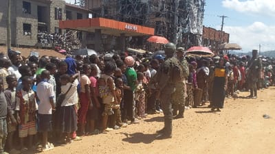 The image shows a large crowd of people lined up in front of a gas station in Kirumba, Democratic Republic of Congo. The gas station is named "ARIKA" and is located in front of a two-story building under construction. The people are waiting in line for gasoline. A soldier in military uniform stands guard at the end of the line. The scene is on a dusty street with a backdrop of hills.