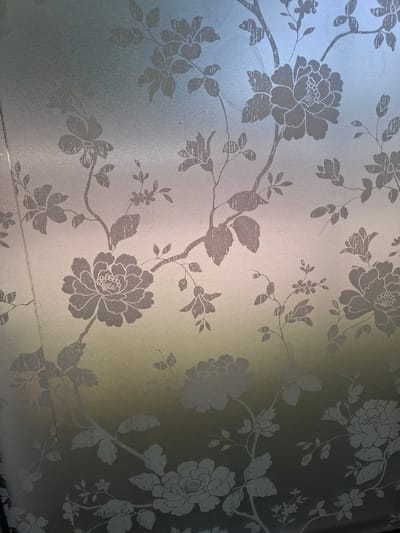 The image shows a close-up of frosted glass with a floral pattern etched onto it. The pattern consists of stylized flowers and leaves in a light gray color. The glass is illuminated from behind, creating a soft glow that highlights the texture of the frosting and the etched design. The lighting creates a gradient effect, with the top of the image appearing lighter and the bottom darker. There are no people or specific landmarks visible in the image that would indicate a location in Uzynagash, Kazakhstan. The image is purely a decorative pattern on glass.