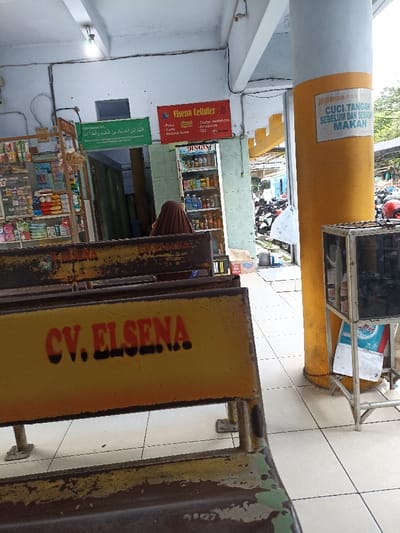 The image shows a close-up of a yellow bench with the words "CV. ELSENA" in red letters. The bench is located under a covered walkway near a large pillar. The pillar is painted yellow and has a sign with the text "MOHON CUCI TANGAN SEBELUM DAN SESUDAH MAKAN". There is a person in a brown hijab sitting on a bench in the background, and a shop with a sign that reads "Elsena Celluler" is visible behind the person. The shop has shelves stocked with various items and a sign that reads "DINGIN" above a cooler. In the distance, there are motorcycles parked near a building. The walkway is tiled and the environment is urban.