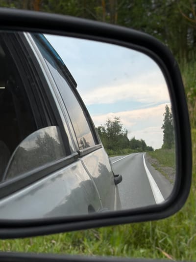 The image shows a side view mirror reflection of a road in Cherdakly, Russia. The road is asphalt with white lines marking the lanes and is surrounded by green grass and trees. The sky is cloudy. The reflection also shows the side of a silver car and the surrounding greenery. The image is taken from a moving vehicle, as evidenced by the blur of the surrounding environment.
