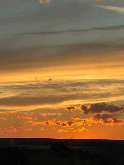 This image depicts a twilight sky over a rural landscape. The sky is filled with layers of clouds, with the lower clouds illuminated in vibrant oranges and yellows, transitioning to lighter shades of orange and then pale blue and gray towards the top. The horizon is marked by a dark silhouette of rolling hills and distant trees, with a faint line of power pylons visible in the far distance. The overall impression is a serene sunset or sunrise over an open, undeveloped area, likely outside of a densely populated urban center. The location in the city of Struino, Bulgaria, implies a rural or semi-rural setting within that region.