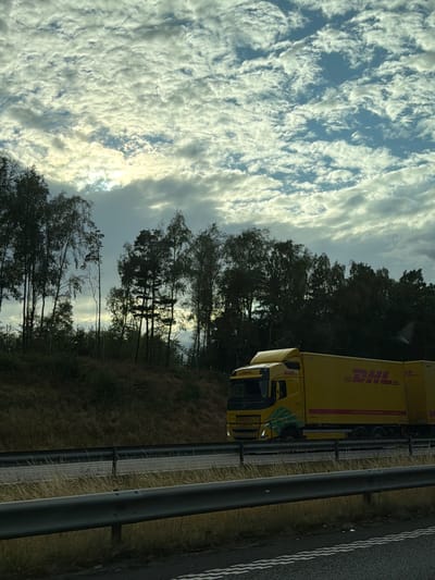 A yellow DHL truck with a red logo is driving on a highway. The truck has a green design on its side. The highway is bordered by trees and dry grass. The sky is cloudy with patches of sunlight. The scene is likely in Åskloster, Sweden, as indicated by the context.