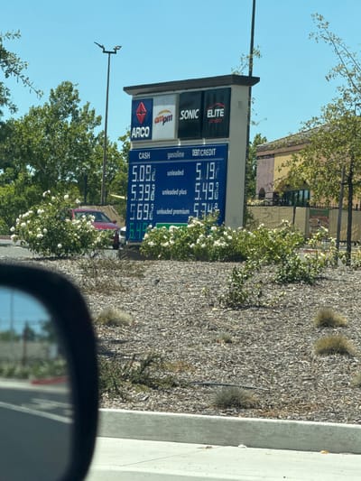 This media file shows a gas station price sign in Citrus Heights, California, on a clear, sunny day. The sign displays prices for gasoline, with different grades of unleaded fuel available. The prices are: Cash $5.09 for unleaded, $5.398 for unleaded plus, and $5.698 for unleaded premium. The debit/credit prices are listed as $5.19, $5.493, and $5.749 respectively.

The gas station appears to be a combined Arco, ampm, and Sonic Drive-In, with a car wash also advertised. The sign has a bright blue background with white text and logos. In the background, there are trees with green leaves and some white flowers blooming. A portion of a building, possibly a restaurant, is visible behind the gas station. The image is taken from the perspective of someone in a car, as a car's side mirror is partially visible in the foreground. The overall scene suggests a typical daytime operation of a busy gas station.