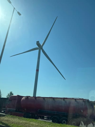 The image depicts a large red tanker truck parked on the side of a road in Jemeppe-sur-Sambre, Belgium. The truck is positioned in the foreground, occupying the lower half of the frame. Behind the truck, a tall wind turbine with its blades angled is visible against a clear blue sky. A lamppost stands to the left of the frame, partially obscured by sunlight. The environment appears to be rural or semi-rural, with a grassy verge in front of the truck and some trees in the distance. The sky is bright and sunny, suggesting daytime.