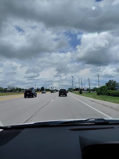 The image is a first-person perspective shot from inside a vehicle traveling on a multi-lane road. The road appears to be in an urban or suburban area, with green trees and some buildings visible on the right side. The sky is overcast with dramatic, dark clouds interspersed with patches of blue sky.

Several vehicles are on the road. A black SUV is in the foreground on the left lane, and a black pickup truck is in the right lane ahead of the viewer. Further down the road, other vehicles are visible, and traffic signals are present. Yellow barrier posts line the left side of the road. A cell tower is visible in the distance on the left, and a sign with "2964" and "US" is visible on the right side of the road. The dashboard of the vehicle from which the photo is taken is partially visible at the bottom of the frame. The scene appears to be within Georgetown, United States of America.