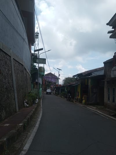 A slightly uphill, narrow asphalt road in Ban Phouviengnoy, Laos, is lined with modest structures. On the left, a tall building with a textured wall and a metallic structure resembling a minaret tower dominates the scene. Garbage bags are visible along the sidewalk. On the right, a row of small buildings with varied exteriors, including patches of color, are present. A parked car is visible in the distance, along with at least two motorcycles. The sky is overcast with a mix of gray and blue patches.