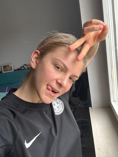 A young, light-skinned individual with blonde hair pulled back from the face is depicted in a close-up, chest-up portrait shot. The setting is an indoor residential space, likely within Roccavione, Italy, with a grey wall in the background to the left and a window to the right. The individual wears a black long-sleeved athletic shirt featuring a white Nike swoosh logo on the left chest and a circular patch on the right chest. This patch contains a white background, a black and white soccer ball, and Cyrillic text, reading "ЯМБОЛ" above the ball and "ФУТБОЛНА МЕЧТА" below it. The person makes direct eye contact with the camera, sticks their tongue out slightly, and has their right hand raised near their forehead, forming a "V" sign (peace sign) with the index and middle fingers. In the background, to the left, a portion of a turquoise-colored headboard or sofa back is visible, behind which sits a small framed picture and a line of colorful toy vehicles. To the right, a white window frame and a light-colored windowsill are present.