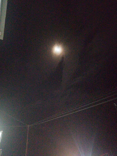 The image depicts a dark, grainy night sky. A bright light source, possibly the moon or a strong artificial light, is visible in the upper central area, casting a soft glow and a subtle shadow. In the lower half of the image, a series of parallel wires stretch across the frame, suggesting the presence of utility poles or a similar infrastructure. A faint light emanates from the bottom edge of the image, possibly from streetlights or vehicles. The overall scene is atmospheric and somewhat indistinct due to the low light conditions and image noise. There are no discernible people or specific landmarks of Gampongpaya, Indonesia in this photograph.