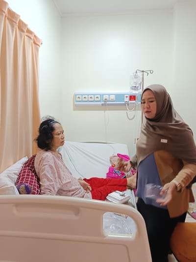 An indoor, medium shot captures a hospital room where an older woman, appearing to be a patient, is propped up in a hospital bed. She is wearing a light pink, patterned pajama top and red pants. Her dark hair is pulled back. Next to her, a woman wearing a hijab and a tan jacket over a blue top stands by the bed. She holds a clear plastic container and appears to be interacting with the patient. The hospital bed is white with an off-white mattress and several pillows. Medical equipment, including an IV stand with a drip bag, is visible in the background, as are electrical outlets on the wall and a light-colored curtain. The room is brightly lit, suggesting daytime. The overall scene implies care and a familial visit.